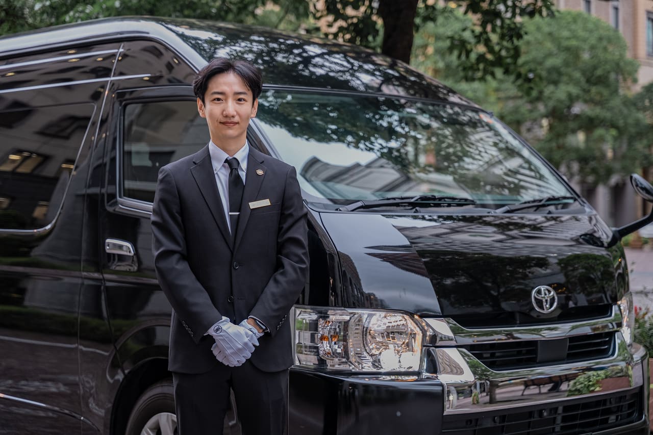 RydAgent's founder in a suit and white gloves beside a black Toyota HiAce