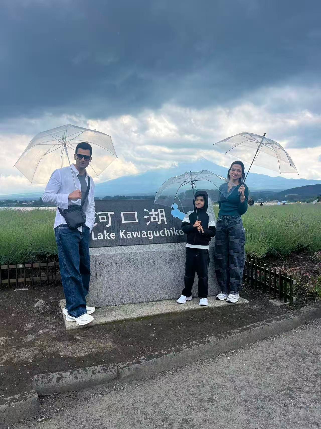 A family of three holding clear umbrellas at the Lake Kawaguchiko marker on an overcast day